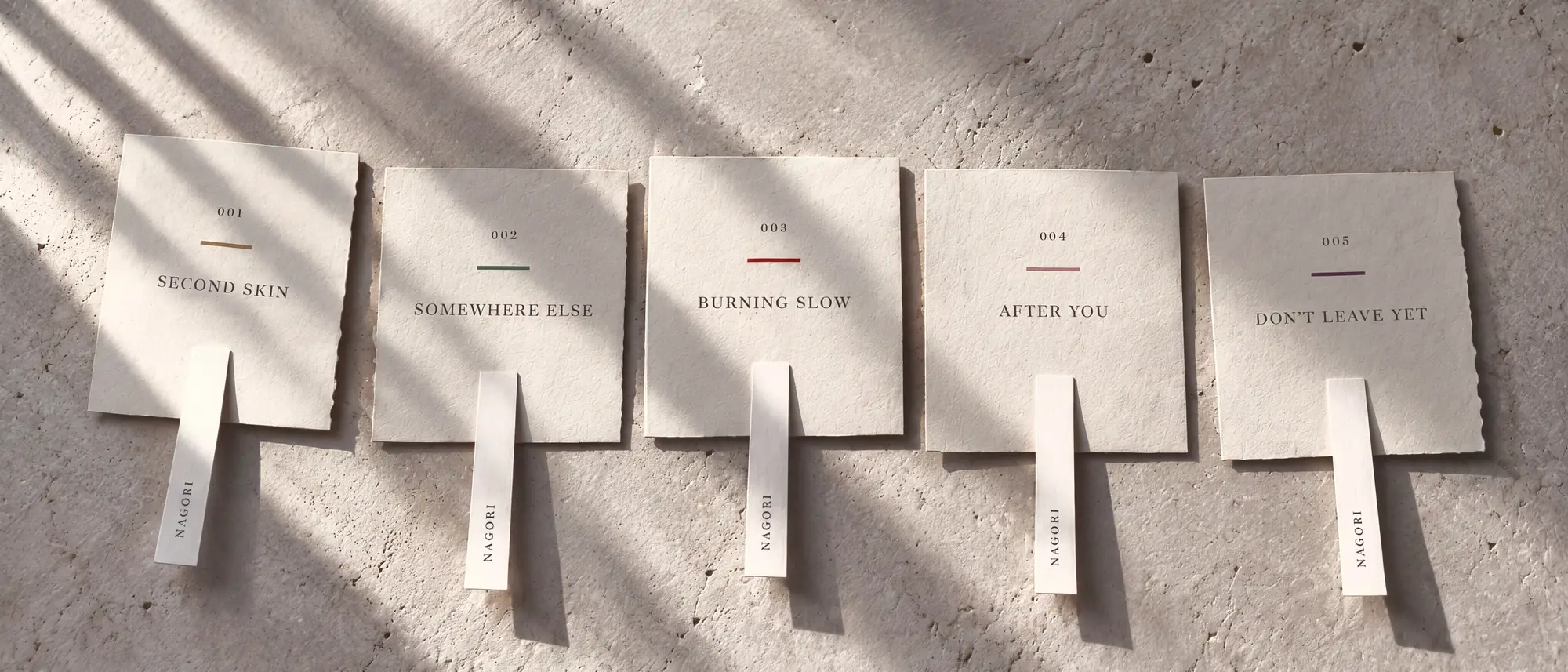 Five deckle-edged cotton-rag cards laid in a row on sunlit concrete, one for each edition &mdash; 001 Second Skin, 002 Somewhere Else, 003 Burning Slow, 004 After You, 005 Don&rsquo;t Leave Yet &mdash; each under its colour rule, with a paper strip marked Nagori below it