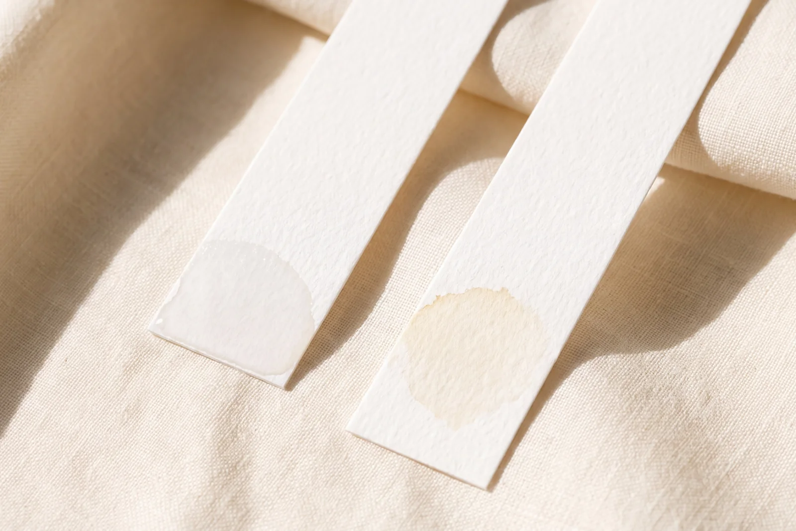 Two paper strips on cream linen: one freshly dipped and still damp, one dipped hours ago with a faint pale-gold mark