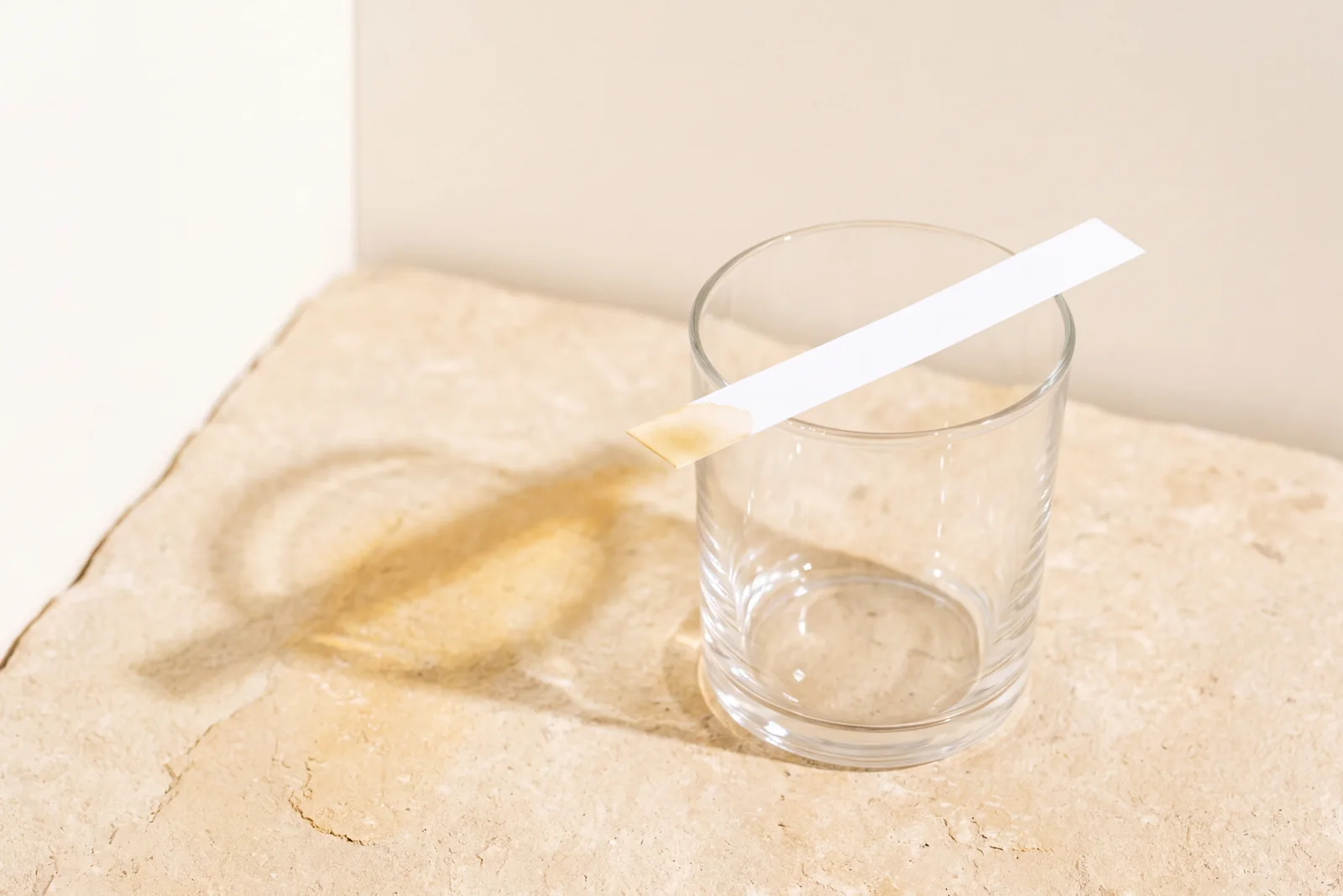 A paper strip resting across a clear glass tumbler on warm stone, its dipped tip faintly stained