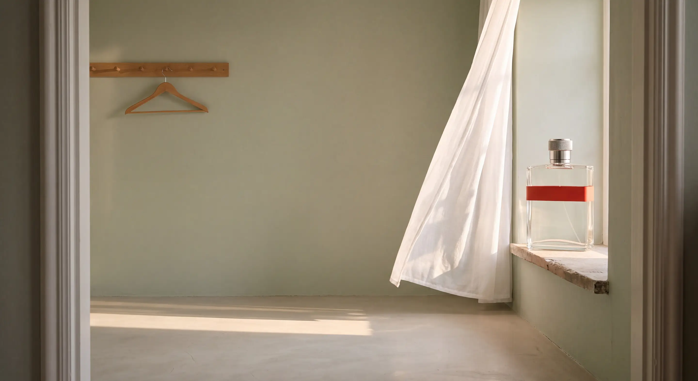 The flask on a stone windowsill in an empty sage-green room, a sheer curtain lifting beside it and one empty hanger on the wall