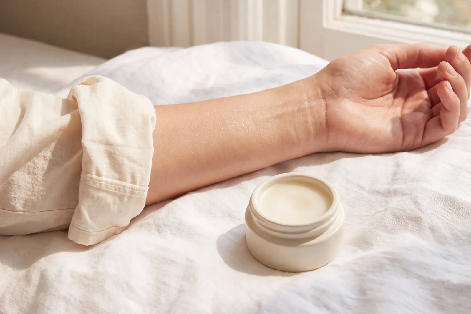 A forearm on white linen in morning sun, a shirt sleeve rolled back, an open pot of balm beside it