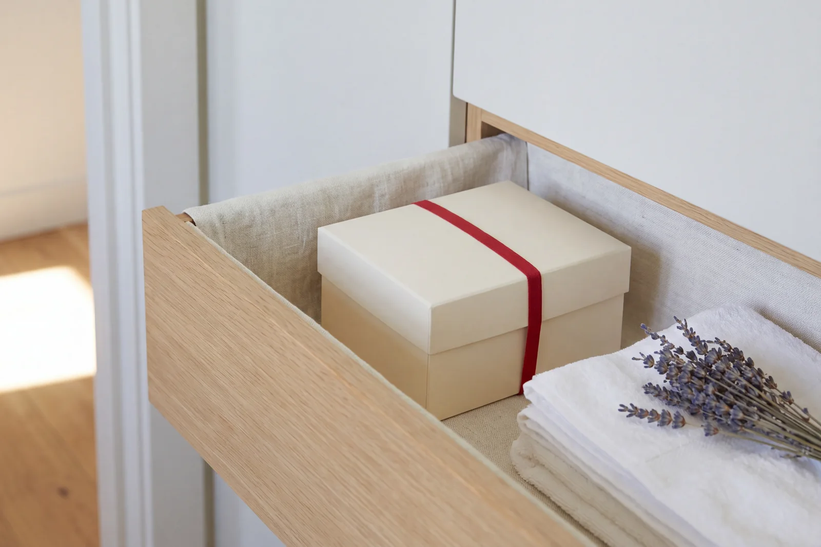 A closed cream box with a red band in a linen-lined drawer, away from the sun on the floor beyond
