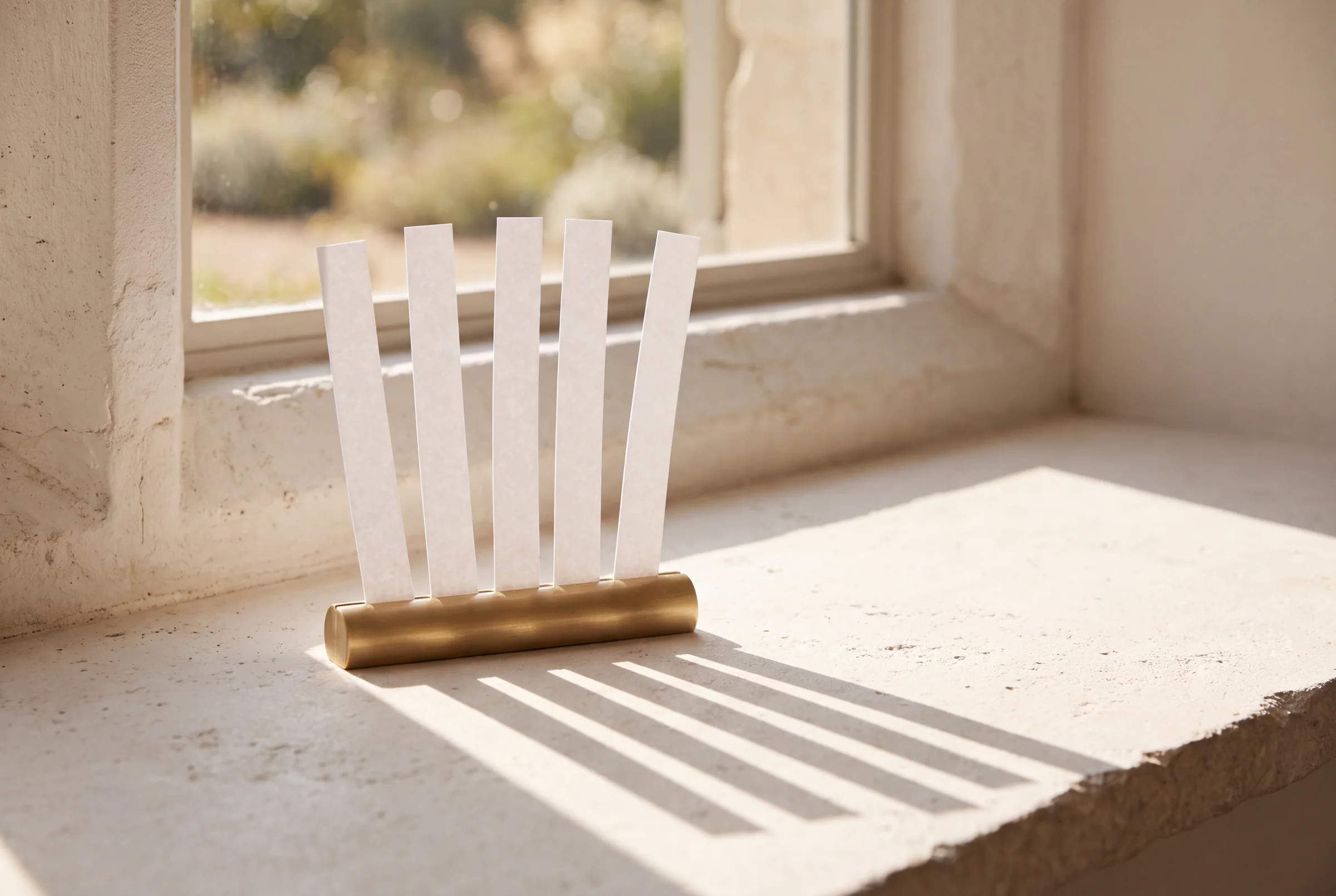 Five paper scent strips standing in a brass holder on a limestone windowsill, five long shadows across the sill